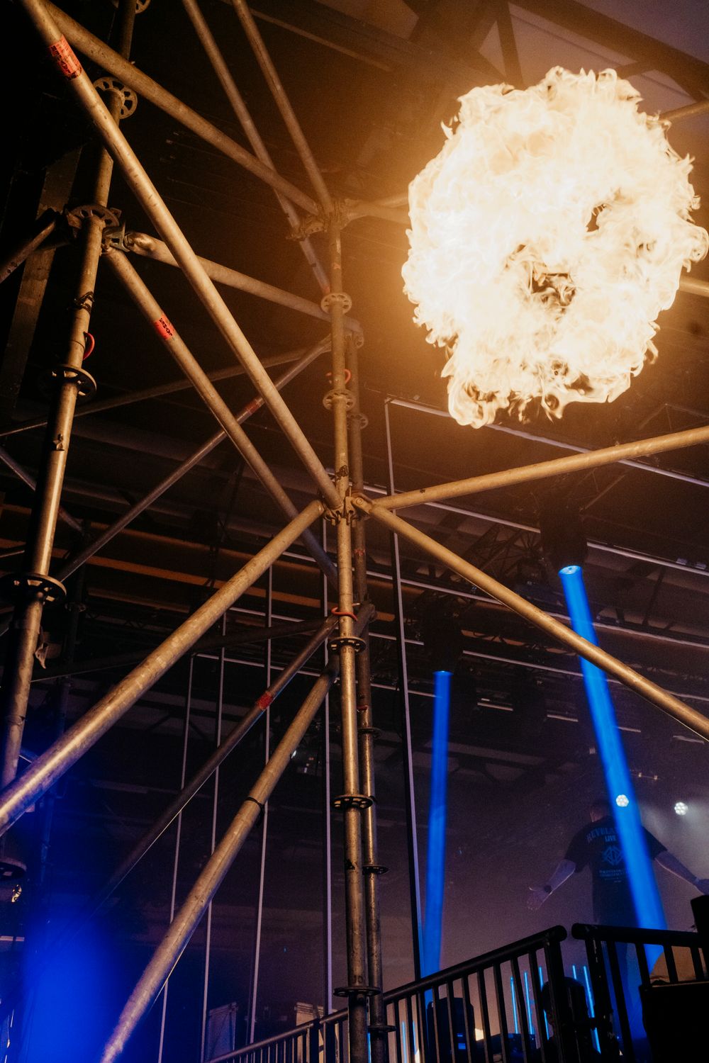 Dramatic fire effects and blue lighting illuminate industrial venue ceiling during live performance.
