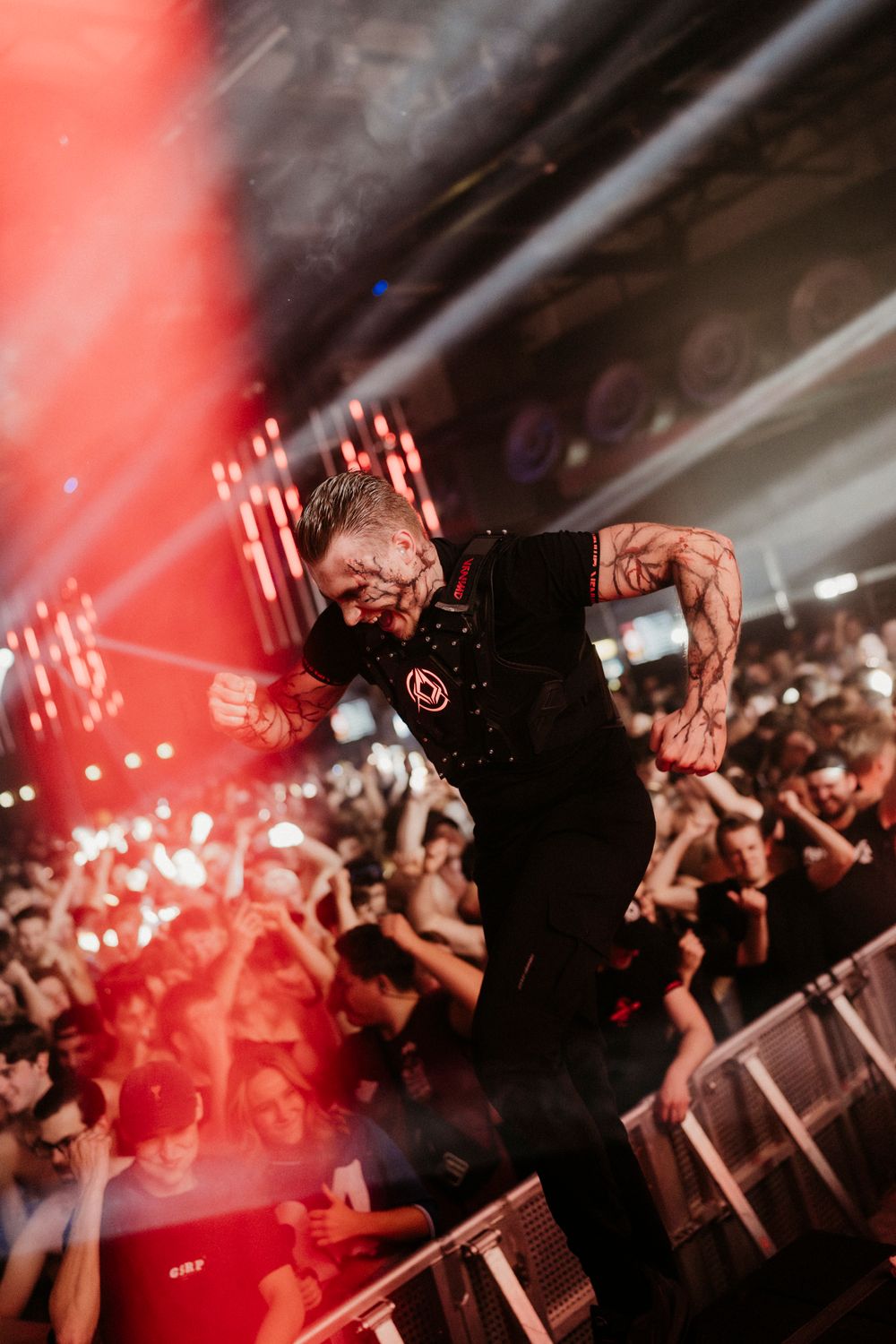 Energetic concert crowd beneath red stage lighting and smoke effects at live music event.