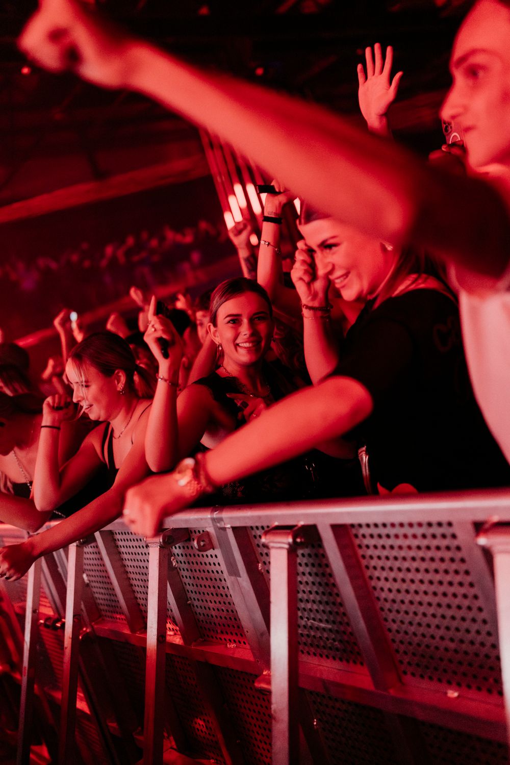 Concert crowd reaching up and cheering at the stage barrier under red lighting.