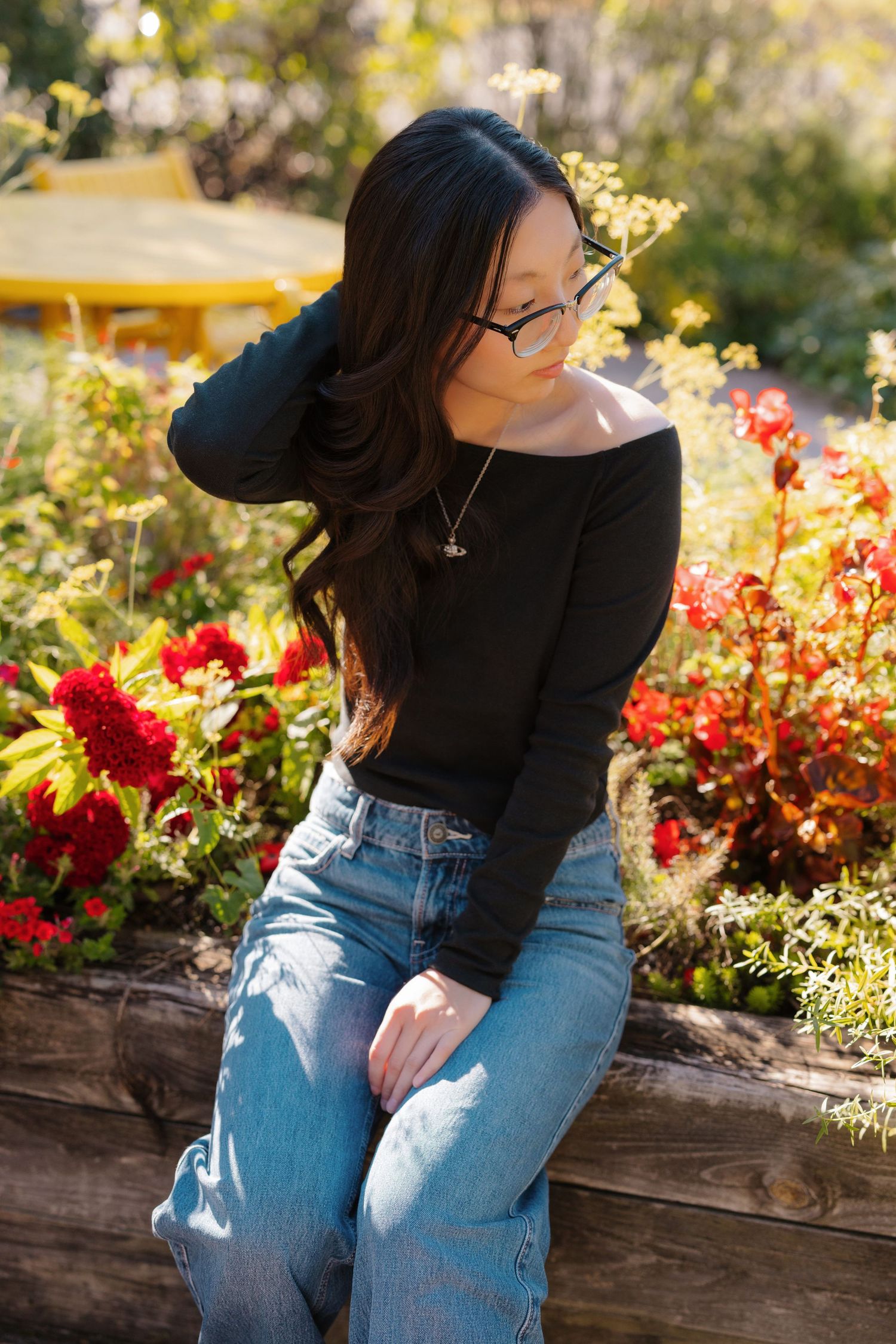Woman in black sweater and jeans poses in garden with vibrant red flowers.