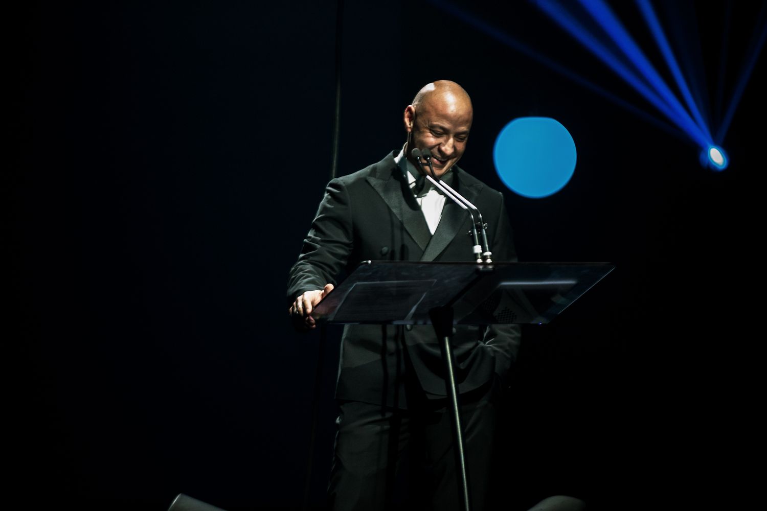 A performer in a black tuxedo stands at a podium on stage with blue spotlights in the background.