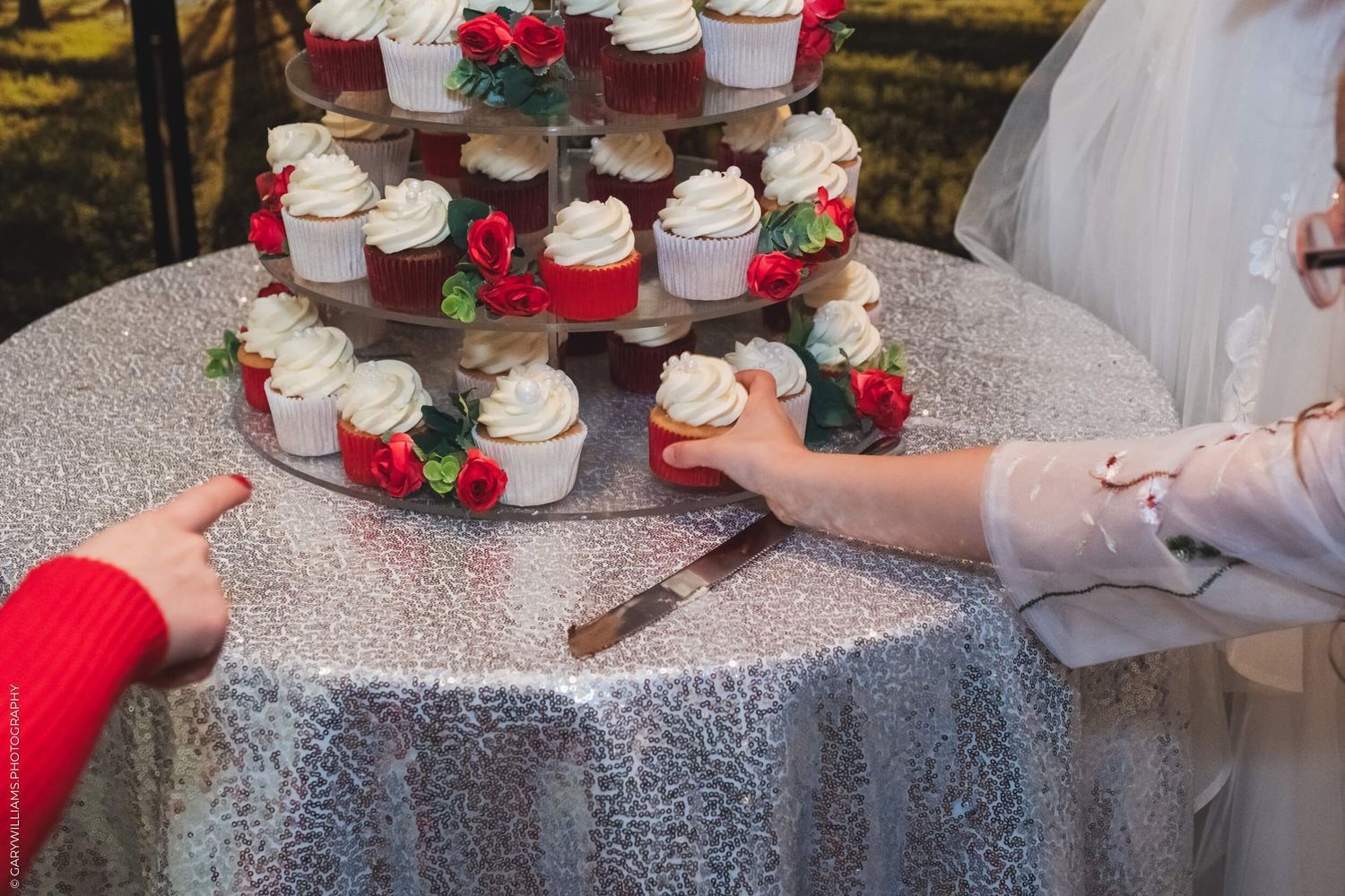 Hands reaching for white frosted cupcakes decorated with red roses on a sparkly silver tablecloth at a wedding reception.