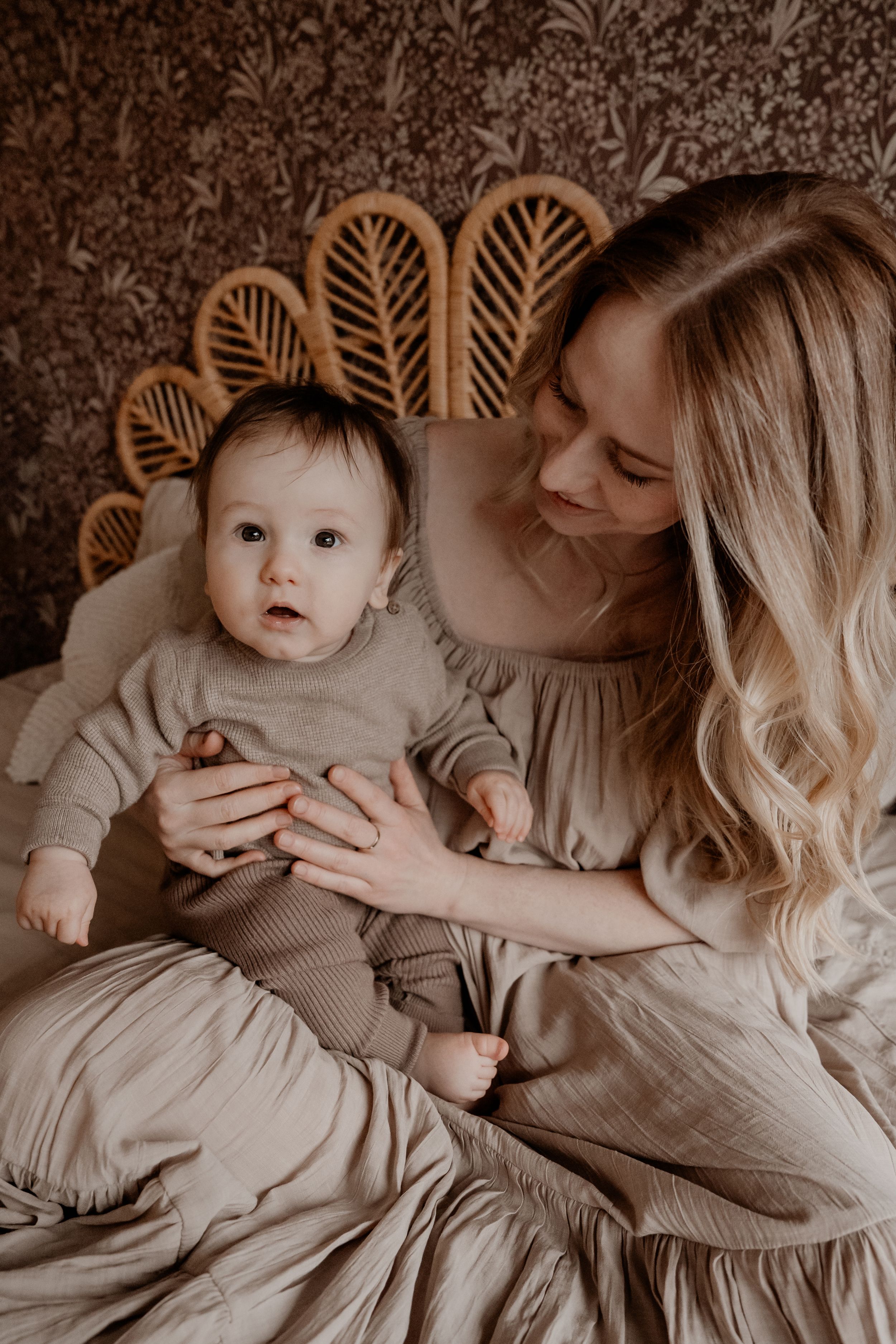 Een teder moment vastgelegd in warme bruintinten, terwijl ouder en baby in bijpassende beige outfits samen zitten bij een rotan hoofdbord.