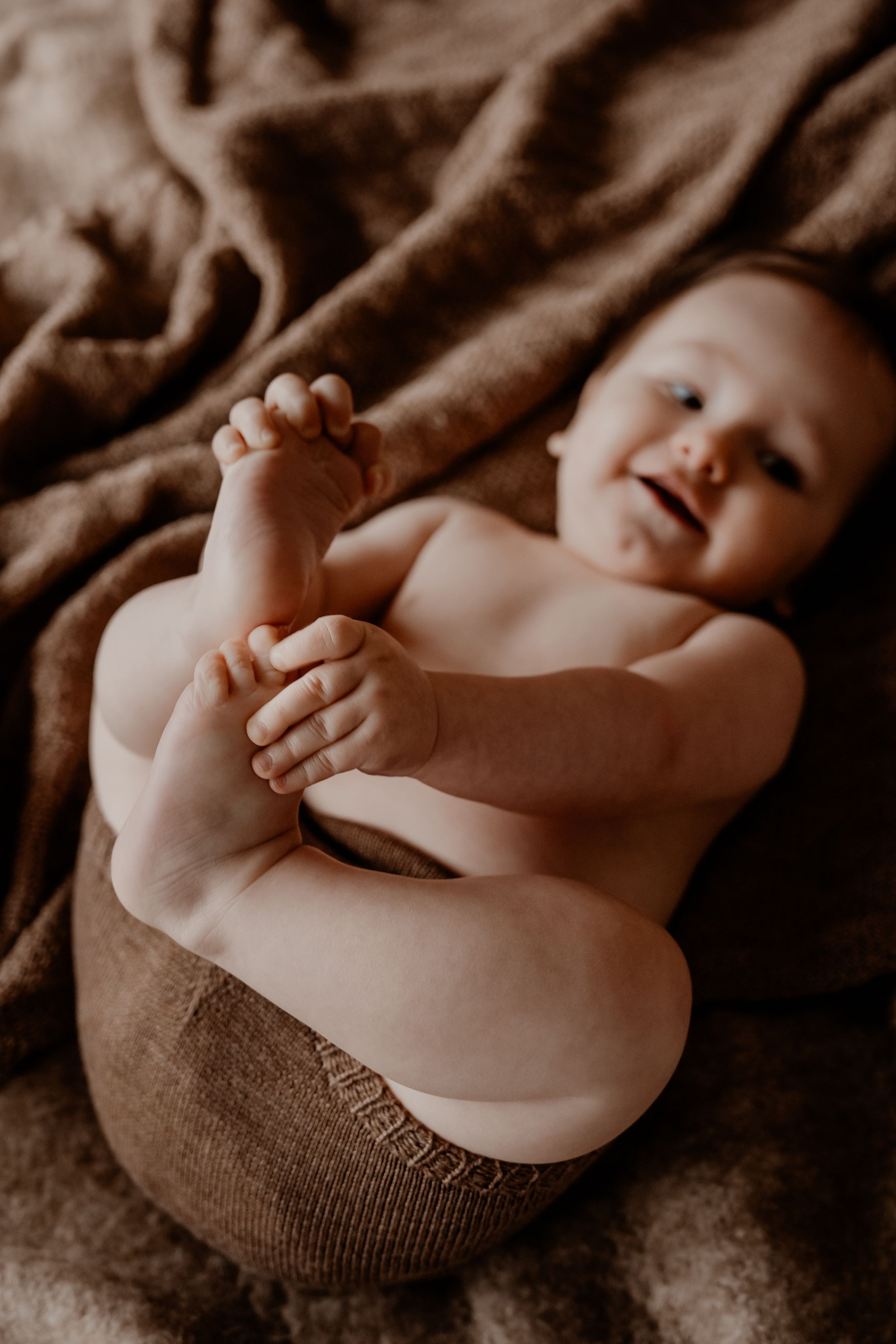 Speelse baby op een bruine deken, reikend naar de teentjes in natuurlijk licht.