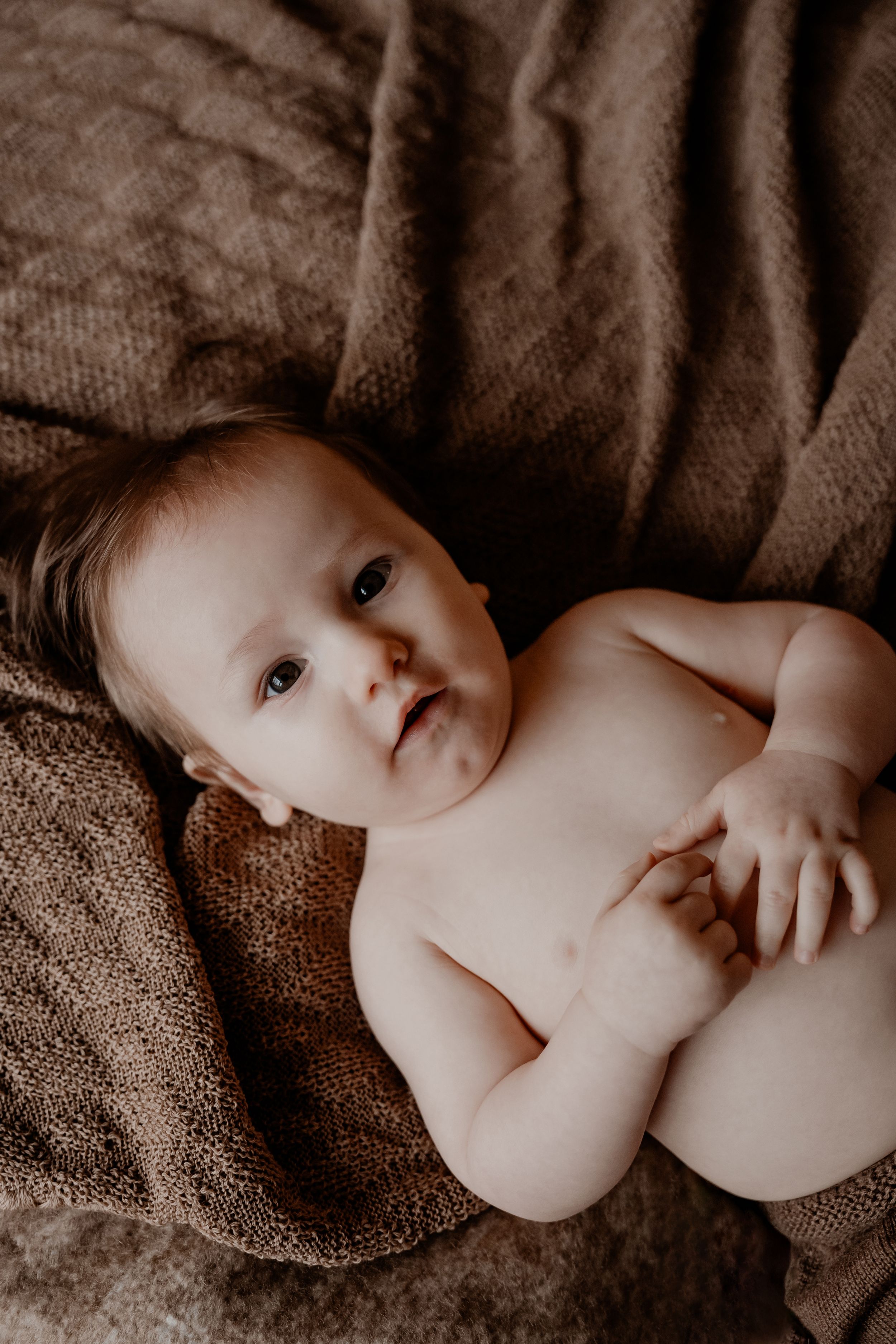 Lieve baby liggend op een textuur bruine deken, met stralende ogen en een rustige uitdrukking.