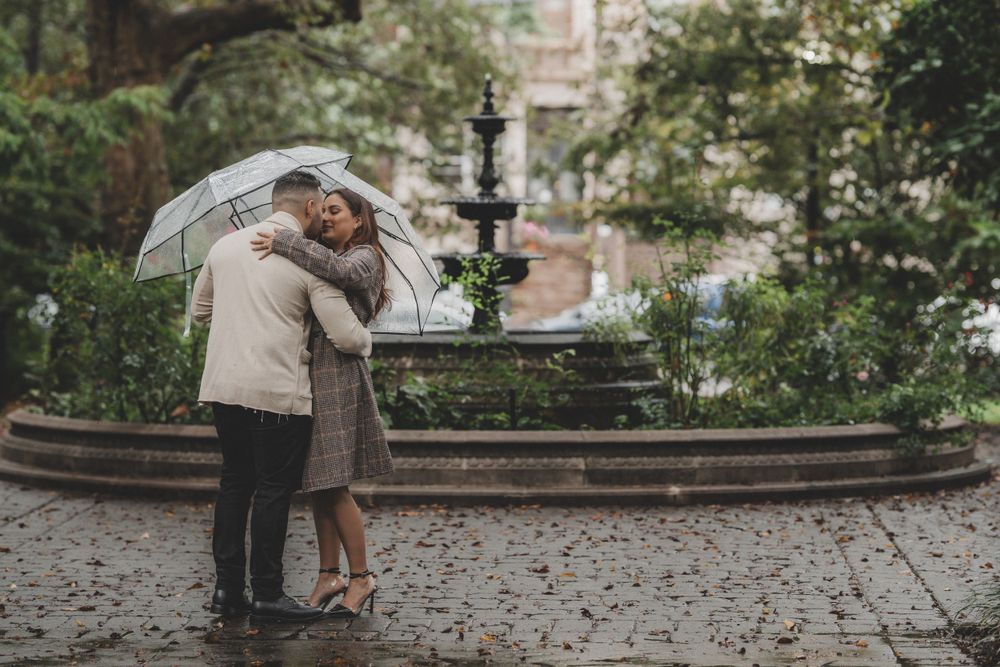 A romantic couple shares an intimate moment under an umbrella near an ornate fountain in a city garden setting.