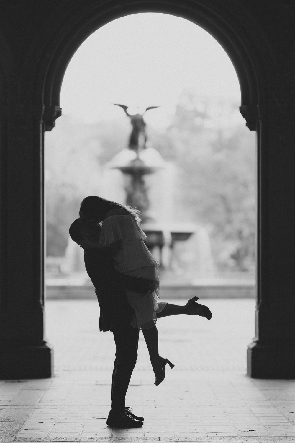 A romantic silhouette of a couple embracing under an archway with a fountain in the background, captured in black and white.