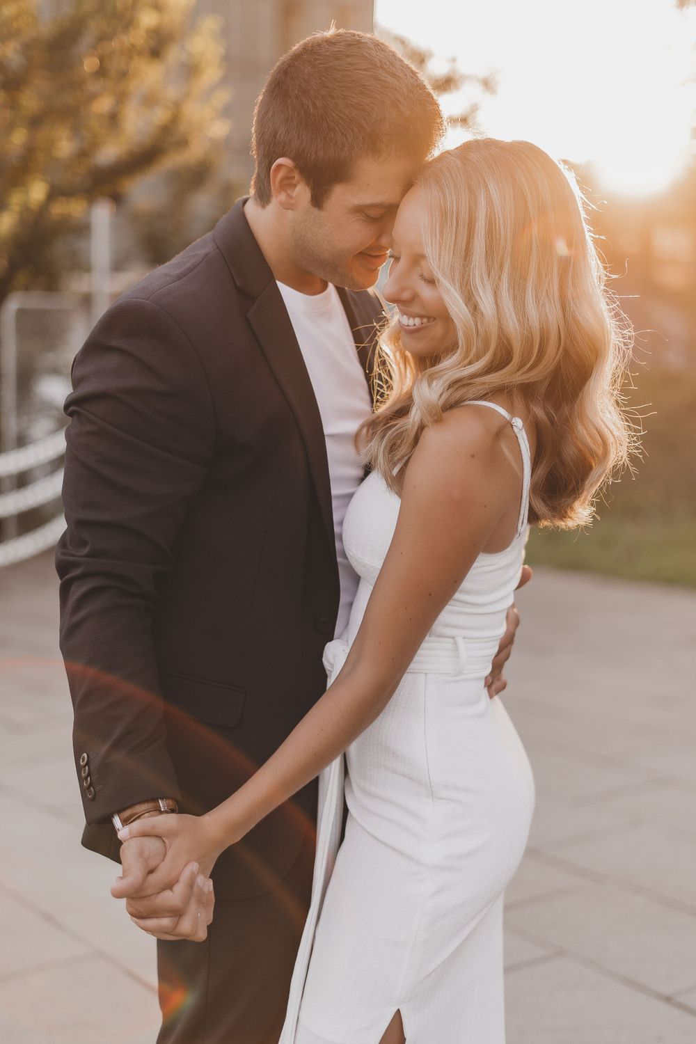 Couple embraces in romantic golden sunset light with her in white dress and him in black suit.