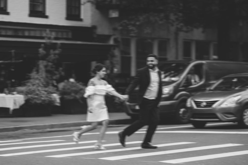 Black and white photo of two people crossing a city street together in an energetic, playful manner.