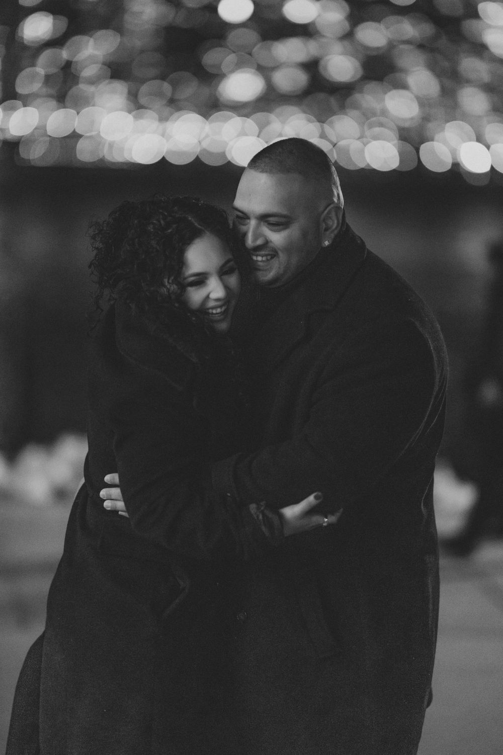 A romantic black and white portrait of a couple embracing outdoors with bokeh lights in the background.