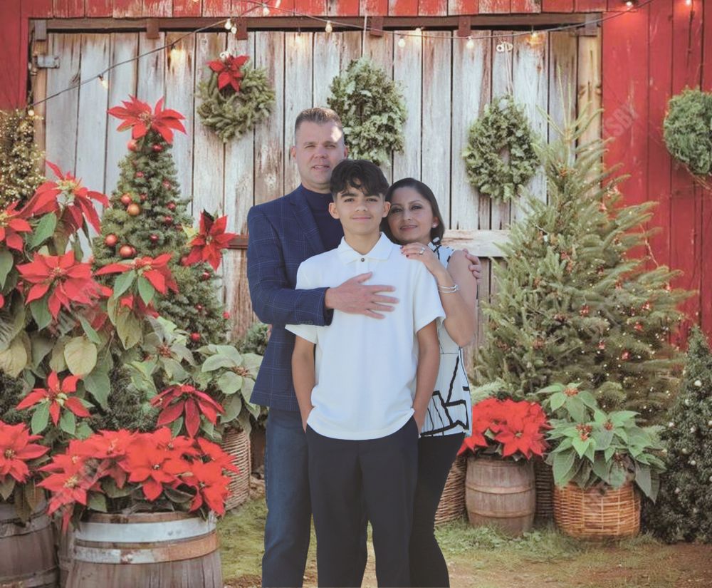 Family of three posing outdoors among Christmas trees, poinsettias, and wreaths on barn wall.