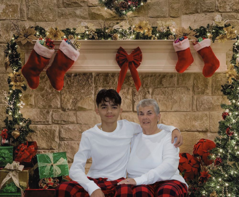 Teen boy and elderly woman in Christmas pajamas posing by decorated fireplace with stockings.