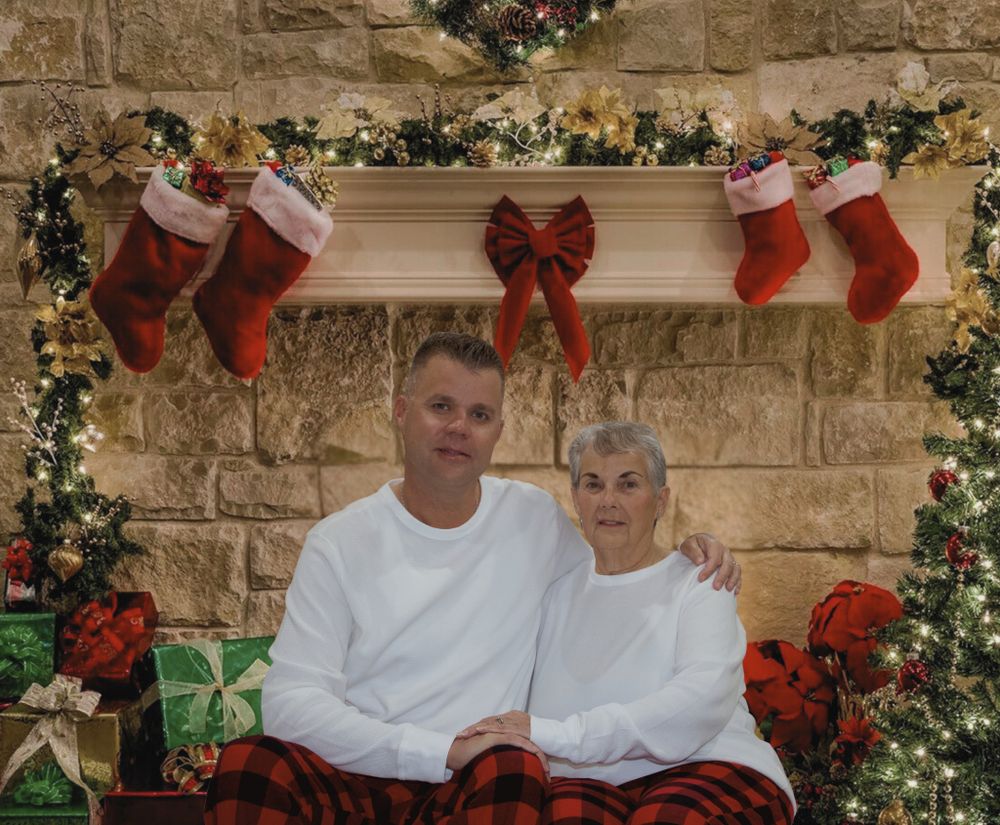Man and elderly woman in white shirts and plaid pants sitting by Christmas fireplace decor.