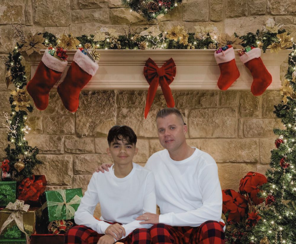 Teen boy and man in white shirts posing together in front of Christmas fireplace decorations.