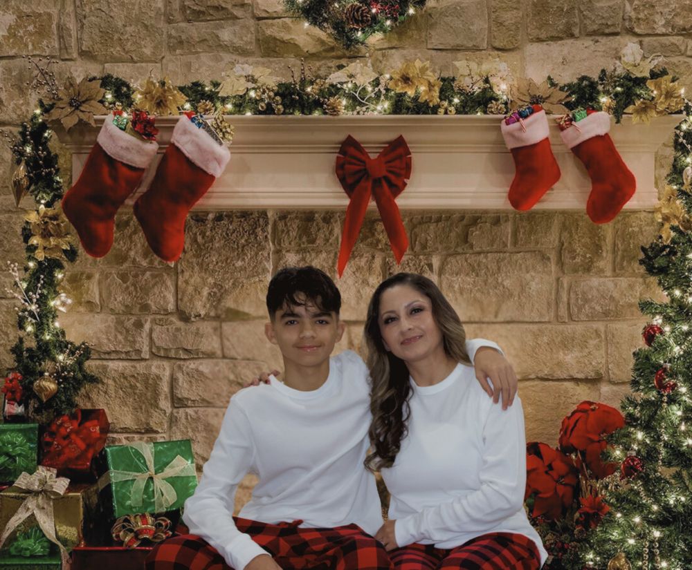 Teen boy and woman in matching Christmas pajamas smiling by decorated holiday fireplace.