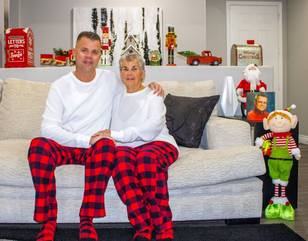 Two adults in red plaid Christmas pajamas sitting on a white couch with holiday decorations.