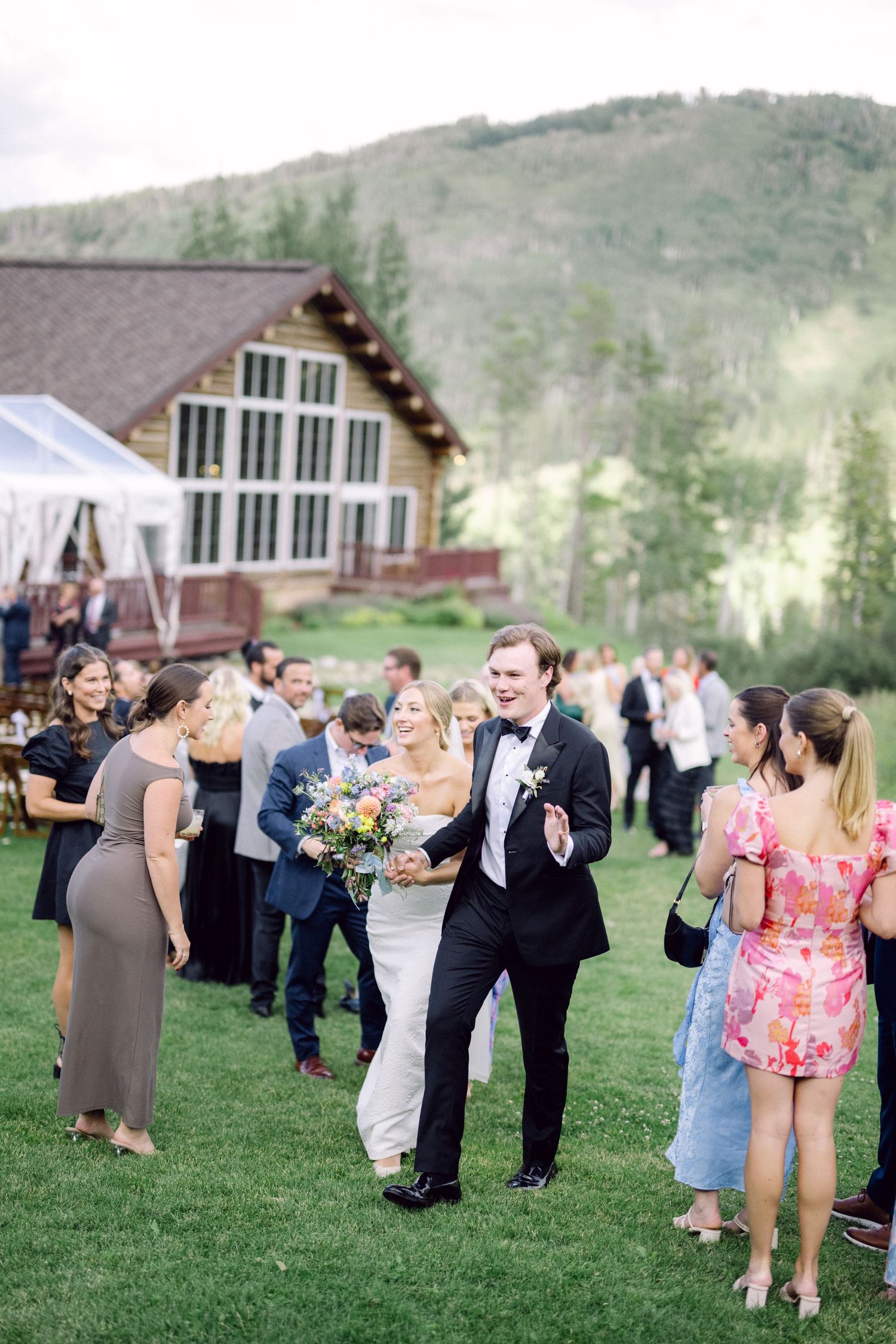Outdoor wedding reception with guests mingling on a lawn near a mountain lodge.