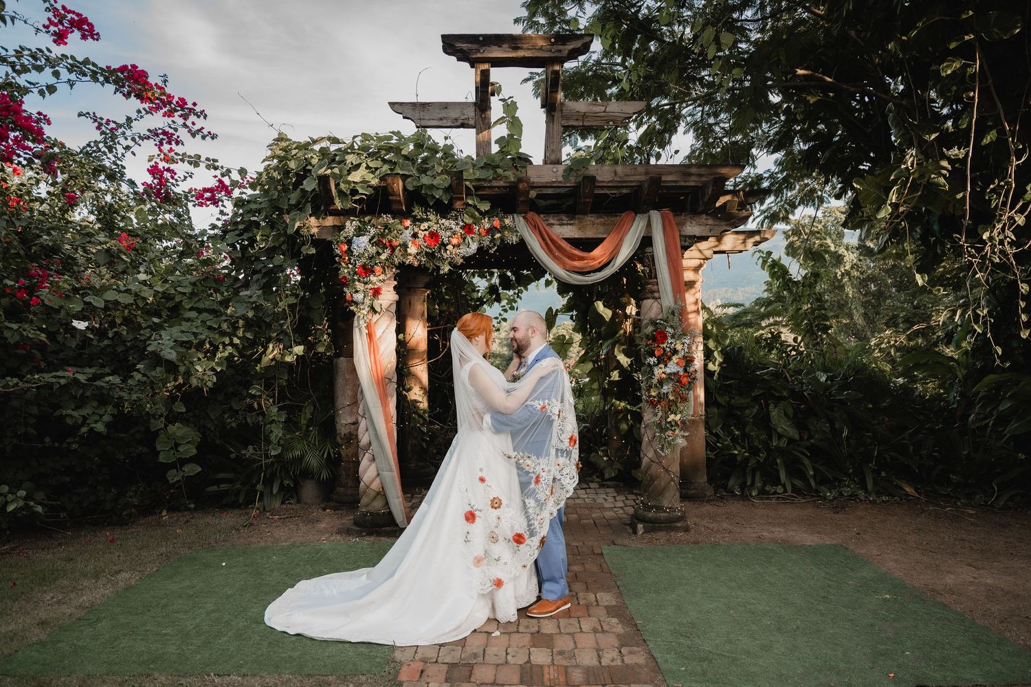 A romantic wedding ceremony under a rustic wooden pergola decorated with orange bunting and floral accents.