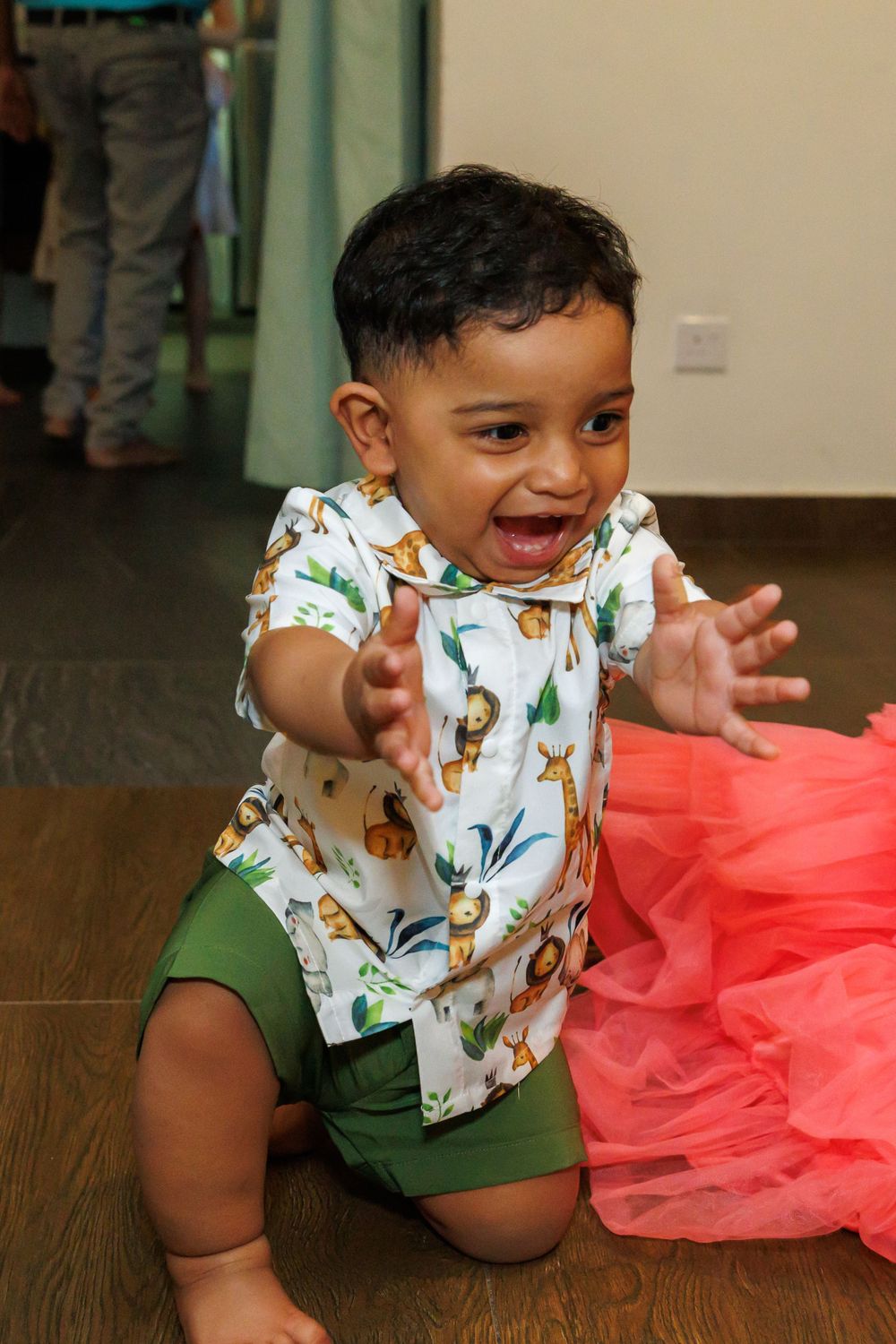 A happy toddler wearing a floral print shirt and green shorts reaches out excitedly.