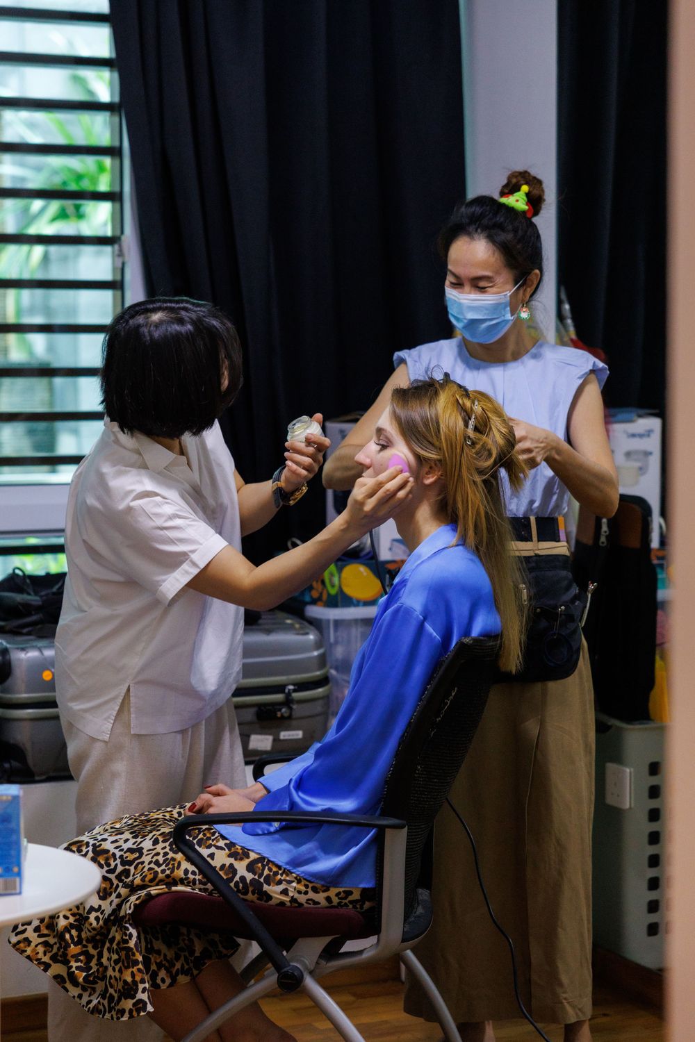 Makeup artists work on styling in a salon setting with protective masks and equipment.