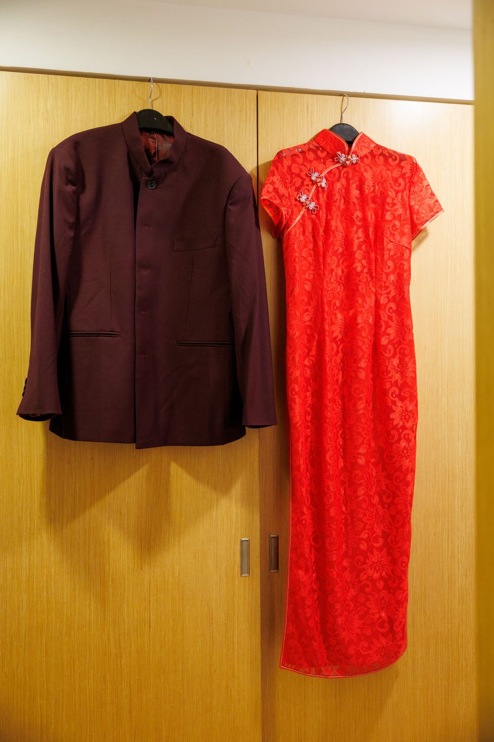 A burgundy suit jacket and red traditional Chinese qipao dress hanging side by side on a wooden wardrobe.