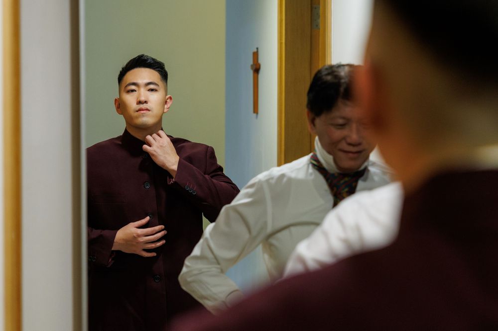 A person in a burgundy sweater adjusts their tie while looking in a mirror, with a doctor in white coat visible behind.