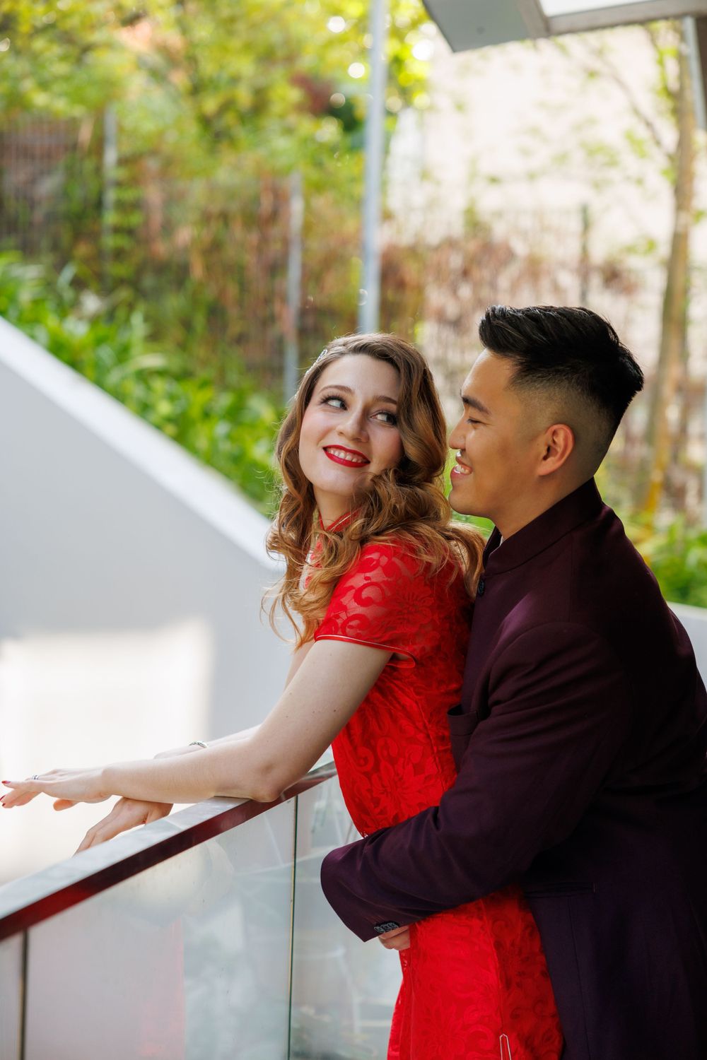 A couple in formal attire share an intimate moment at a white piano, with her in red and him in burgundy.