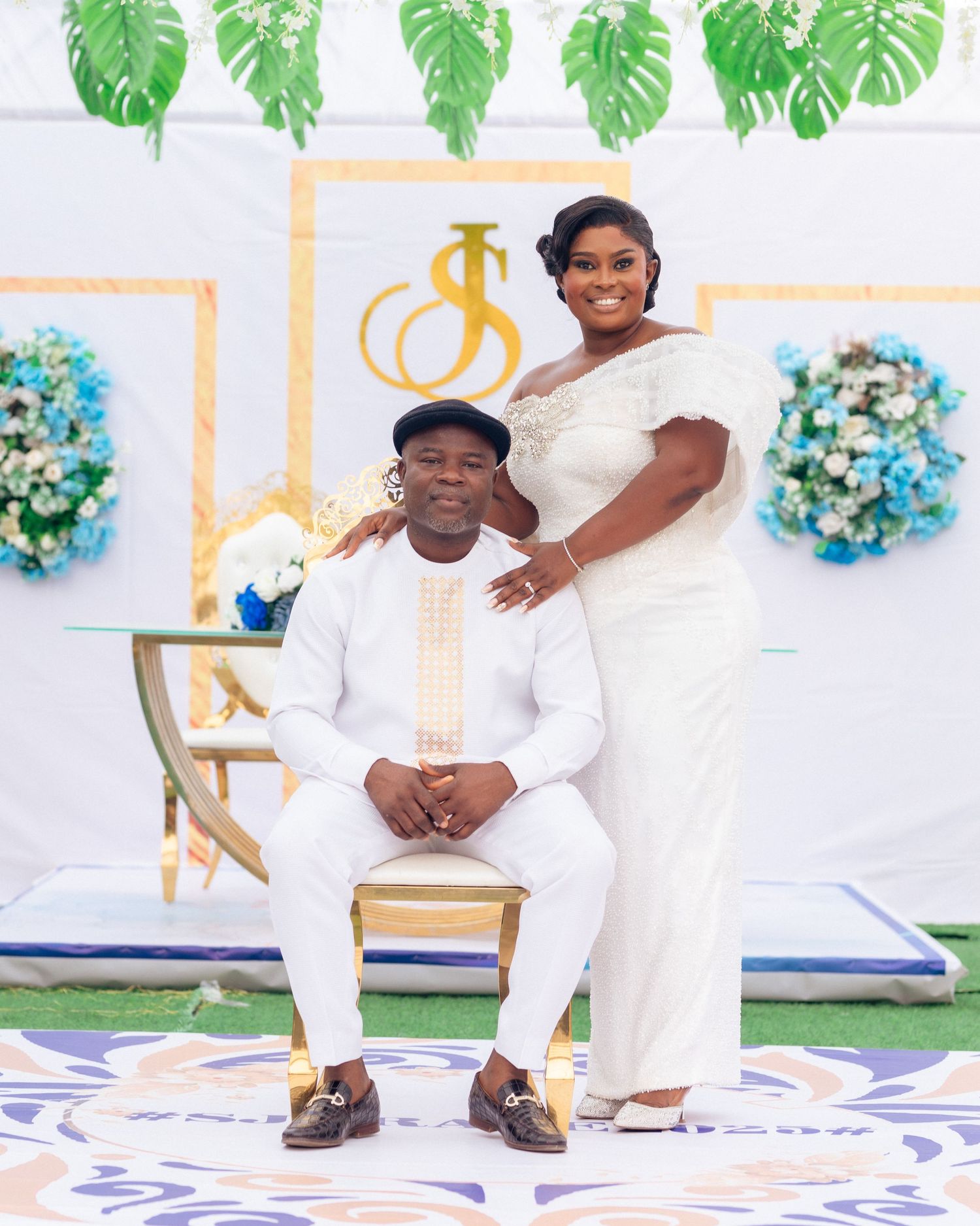 Elegant couple in matching white outfits pose at a formal event with decorative monstera leaves overhead.