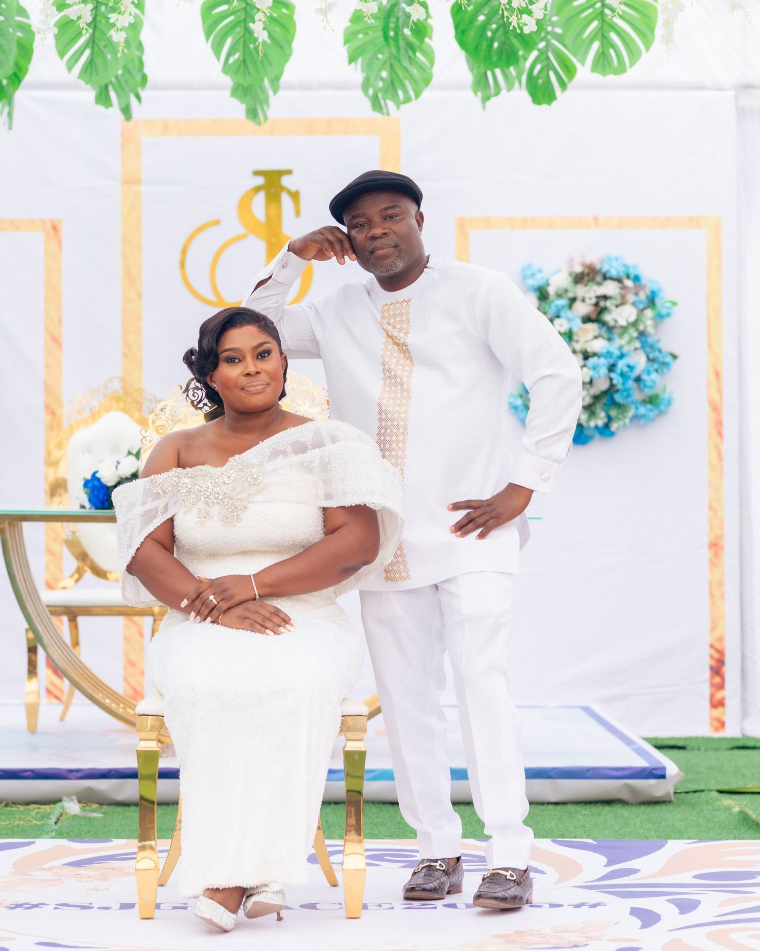 Couple in coordinated white attire poses together at a decorated event space with tropical greenery.