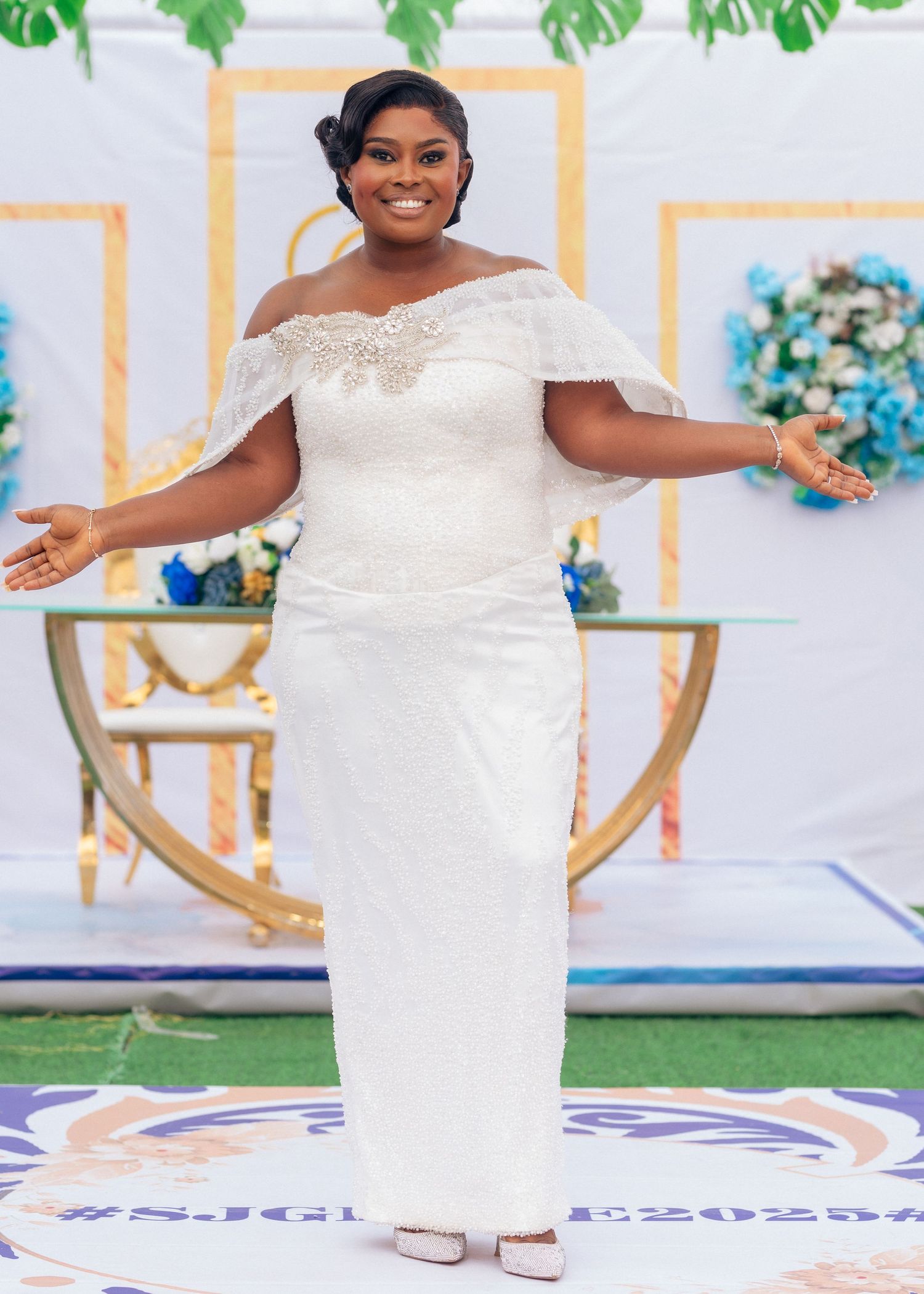 Woman in white off-shoulder gown poses joyfully on a decorative rug with tropical backdrop.