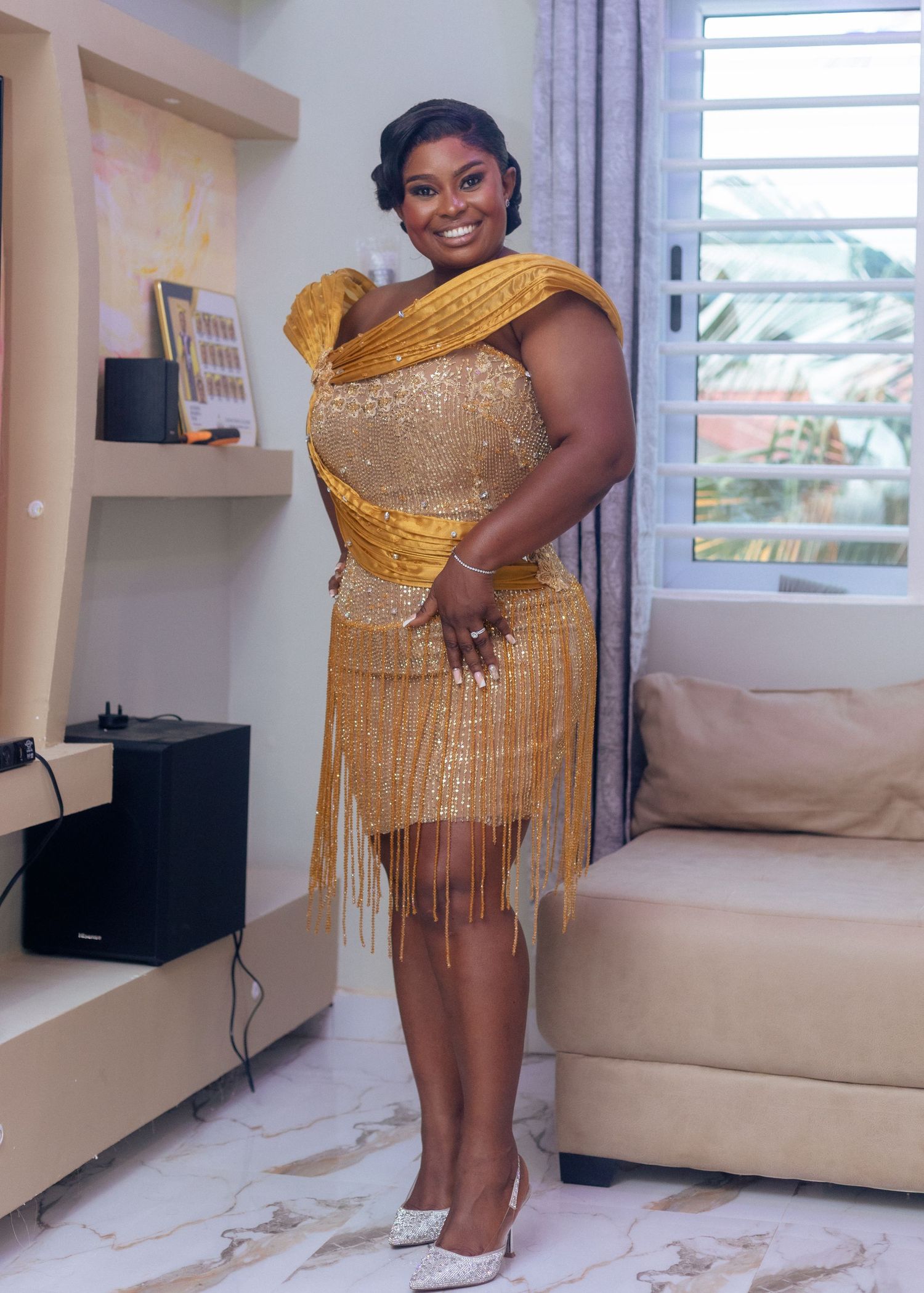 Woman poses in glamorous gold fringe dress with matching scarf in modern interior setting.