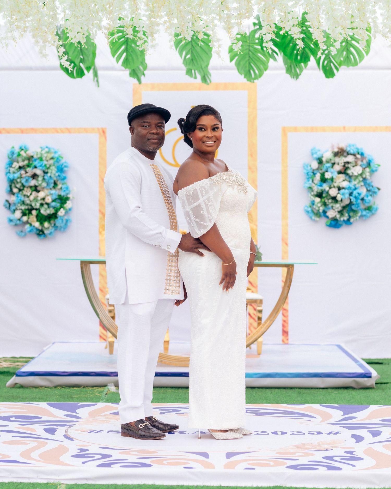 Couple poses together in white outfits against a tropical-themed backdrop with monstera leaves and blue floral decor.