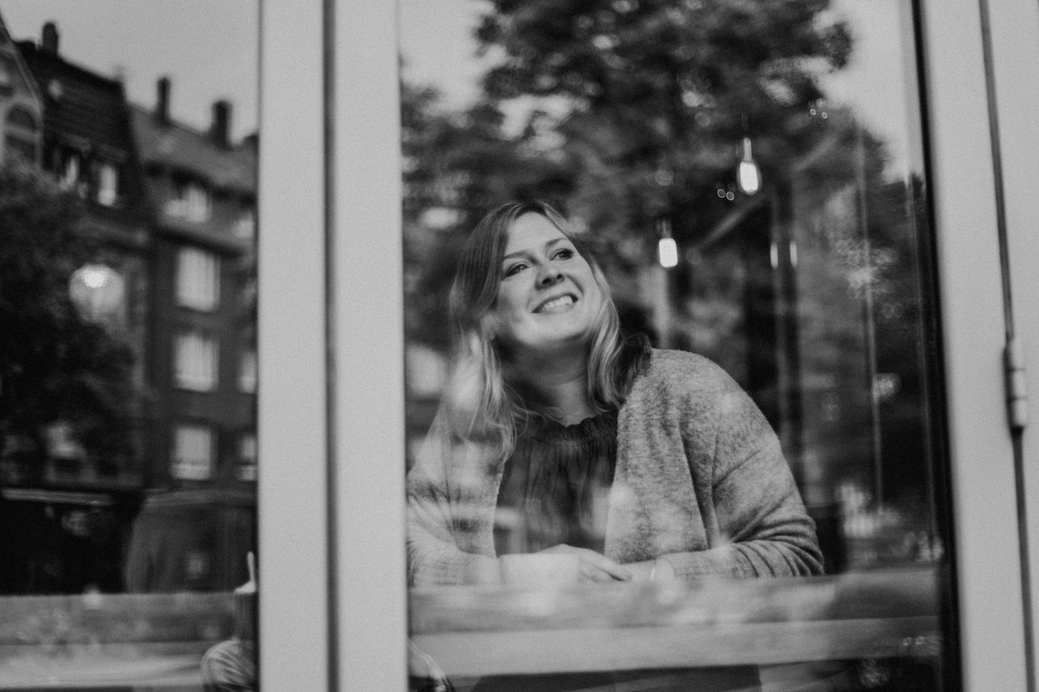 Frau sitzt lächelnd in einem Café, schaut durch ein Fenster. Draußen sind unscharfe Gebäude und Bäume zu sehen. Schwarz-weiß Foto.