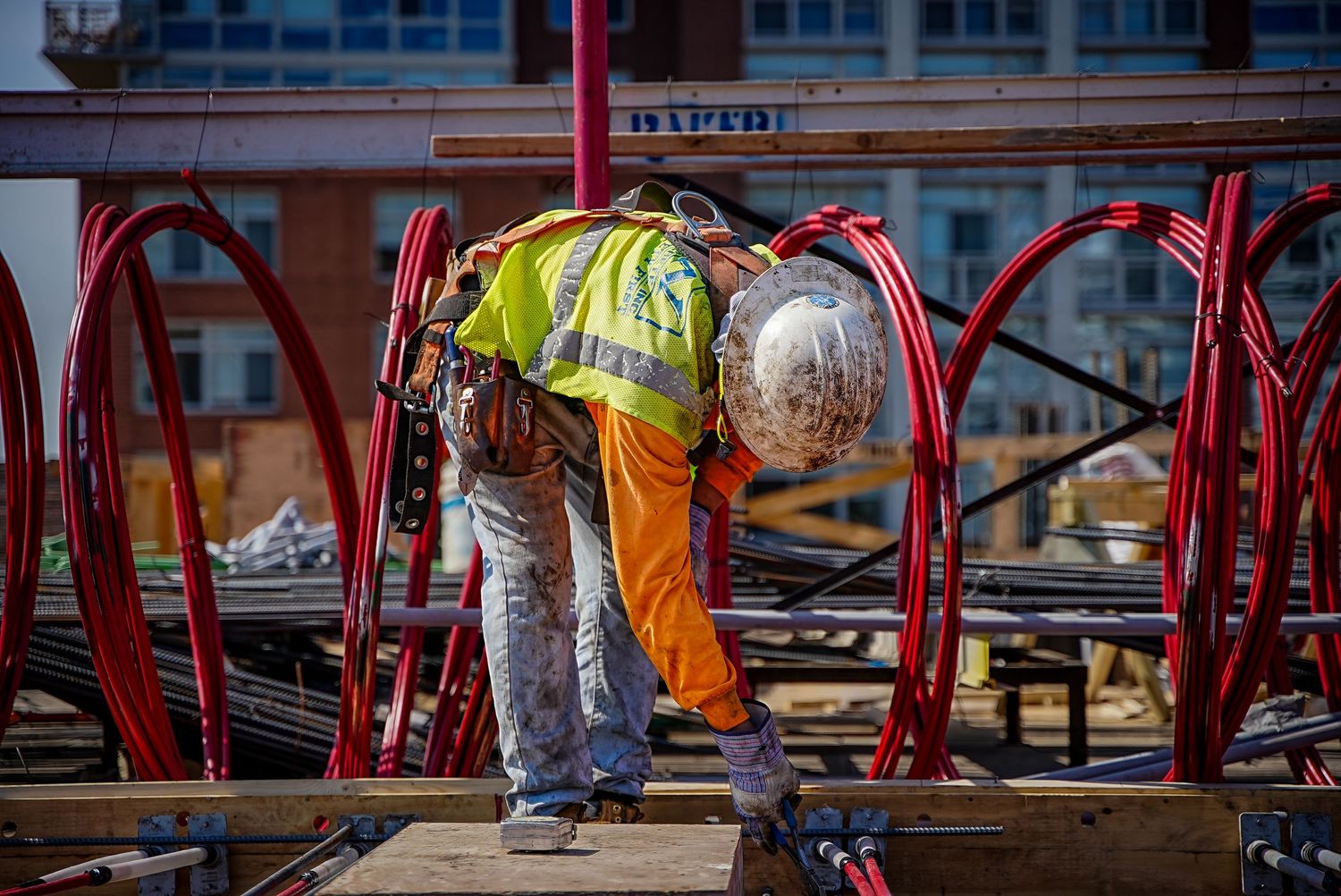 Construction worker in safety vest and hard hat bends over to work on red metal structure at construction site.