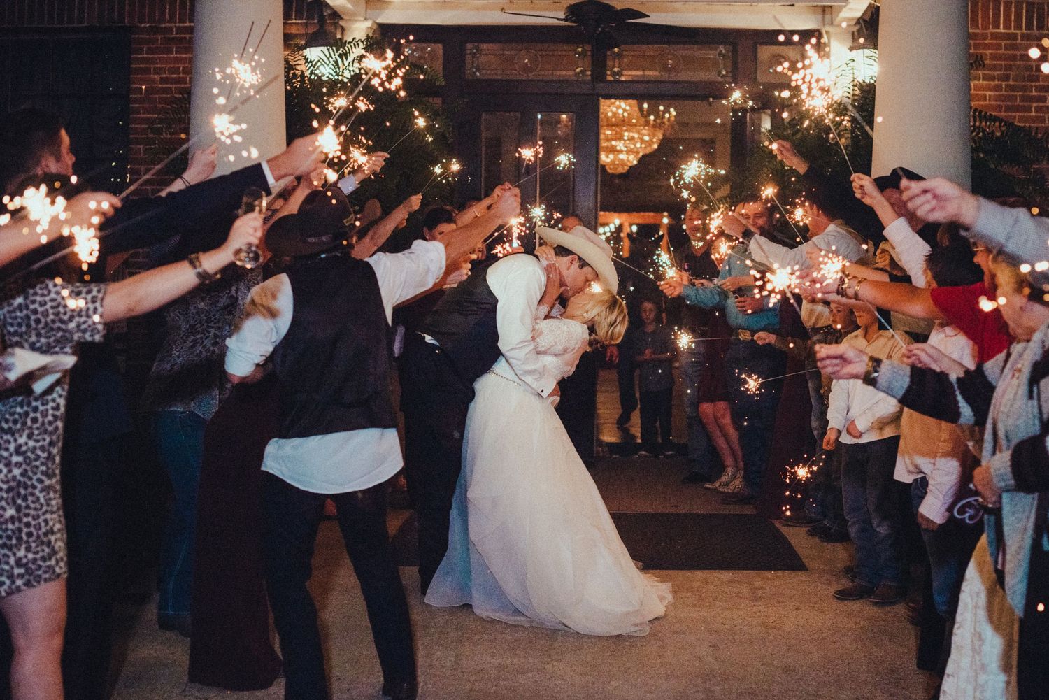 Newlywed couple exits their wedding reception through a tunnel of sparklers held by celebrating guests at night.