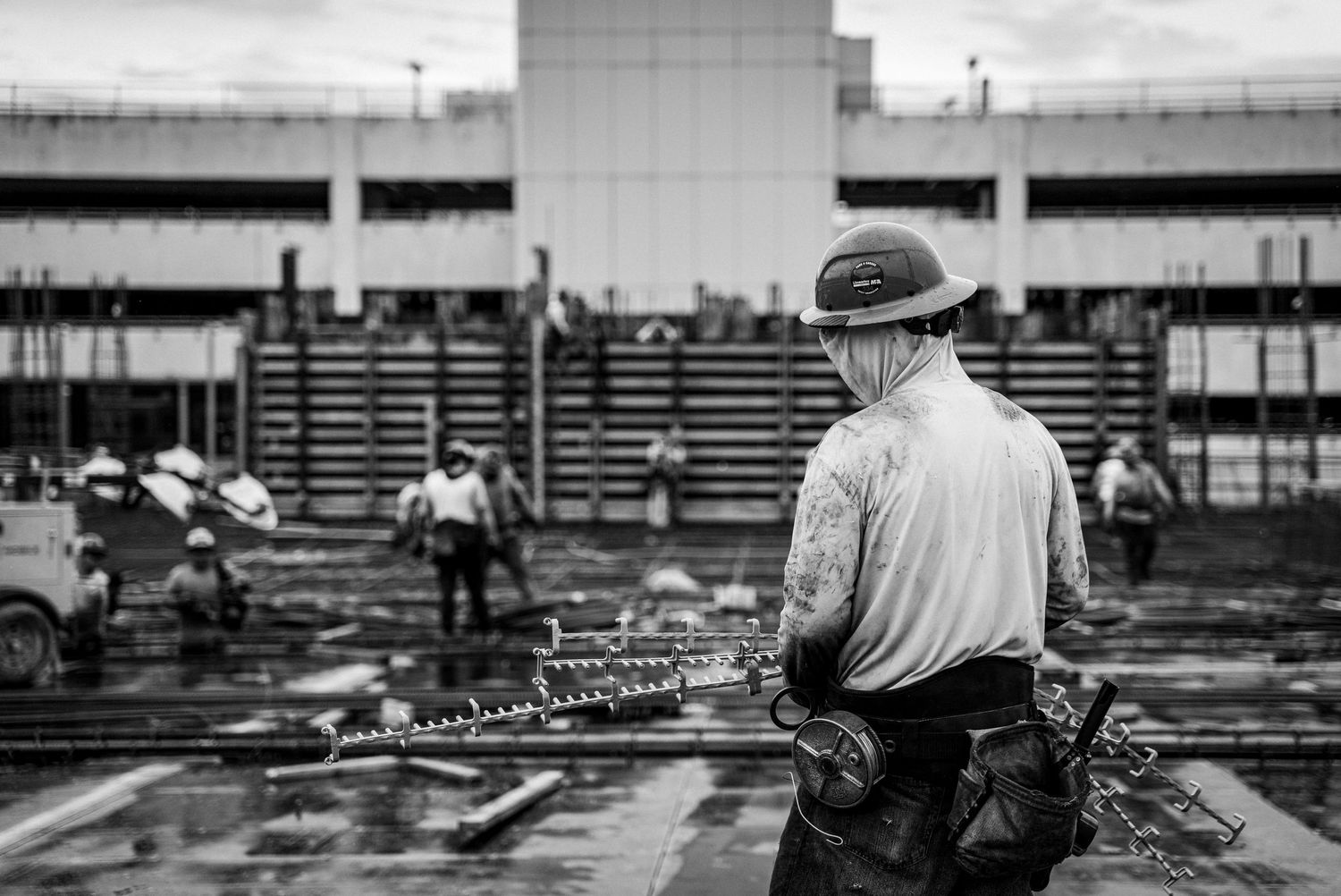 Construction worker in hard hat and safety gear overlooks building site from elevated position in black and white photo.