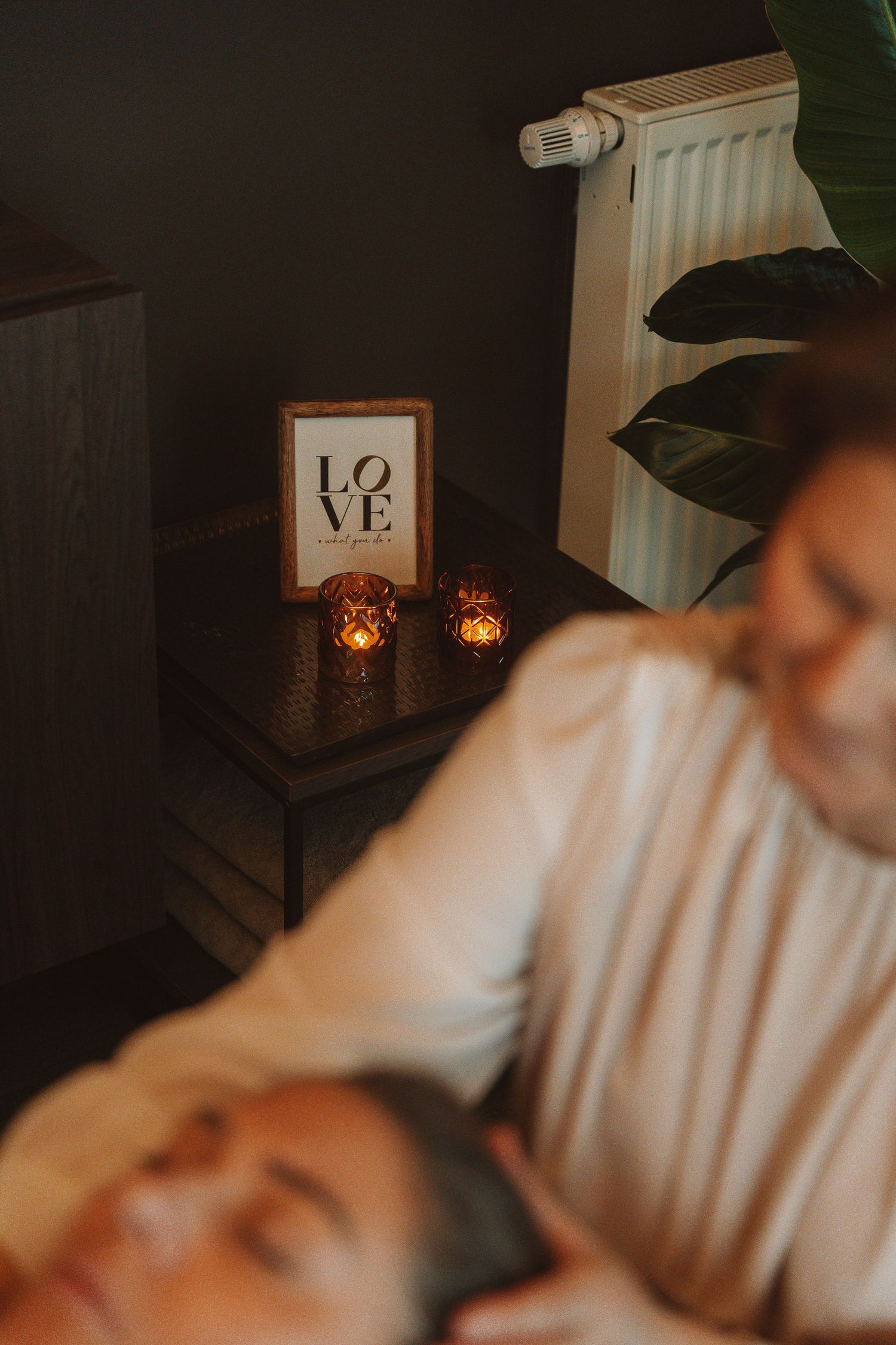 Cozy room interior with lit candles, LOVE sign artwork, and radiator by houseplant in warm atmospheric lighting.