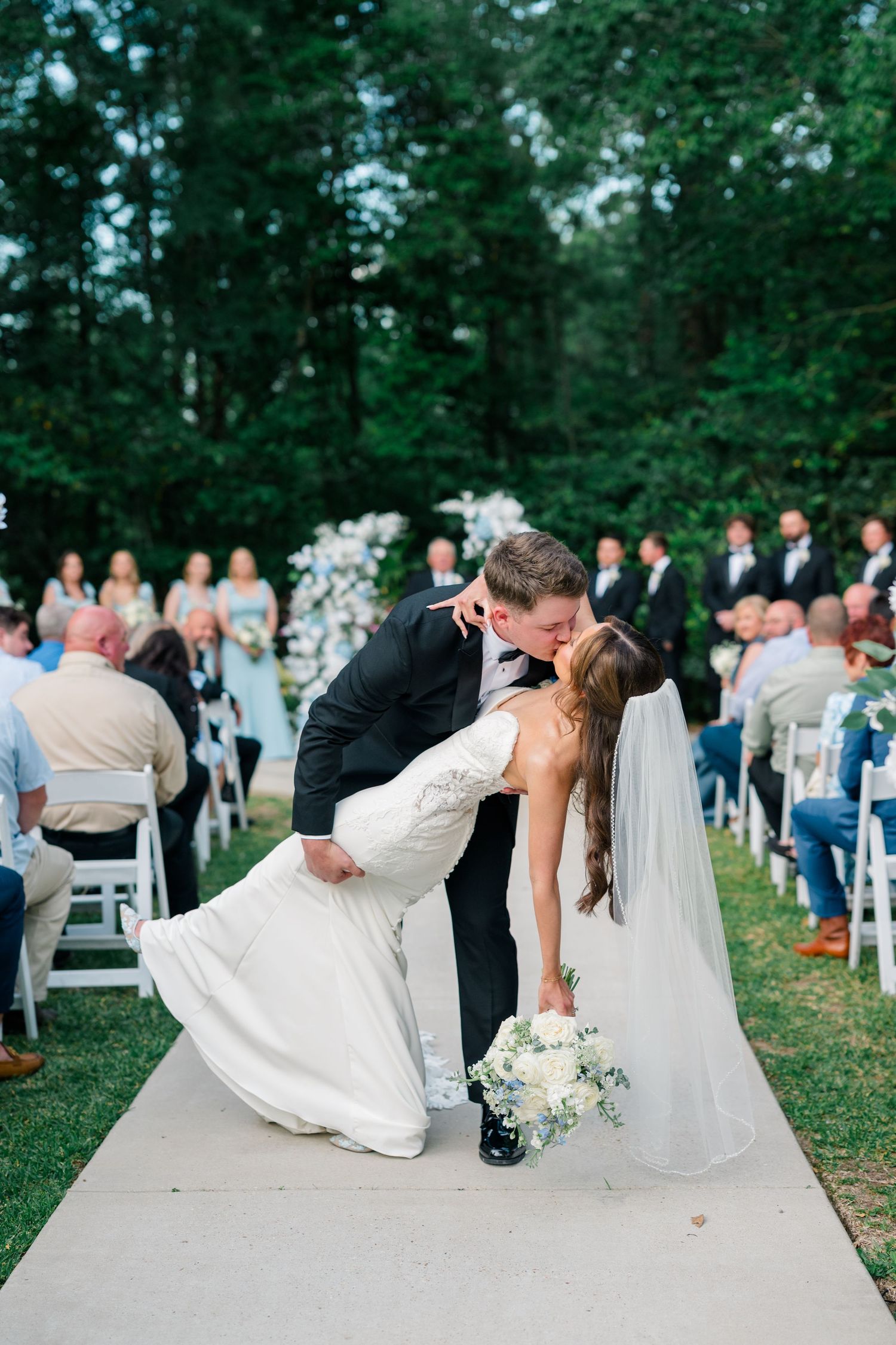 Close-up wedding portrait of newlyweds embracing with white veil and blue shirt against blurred outdoor background.
