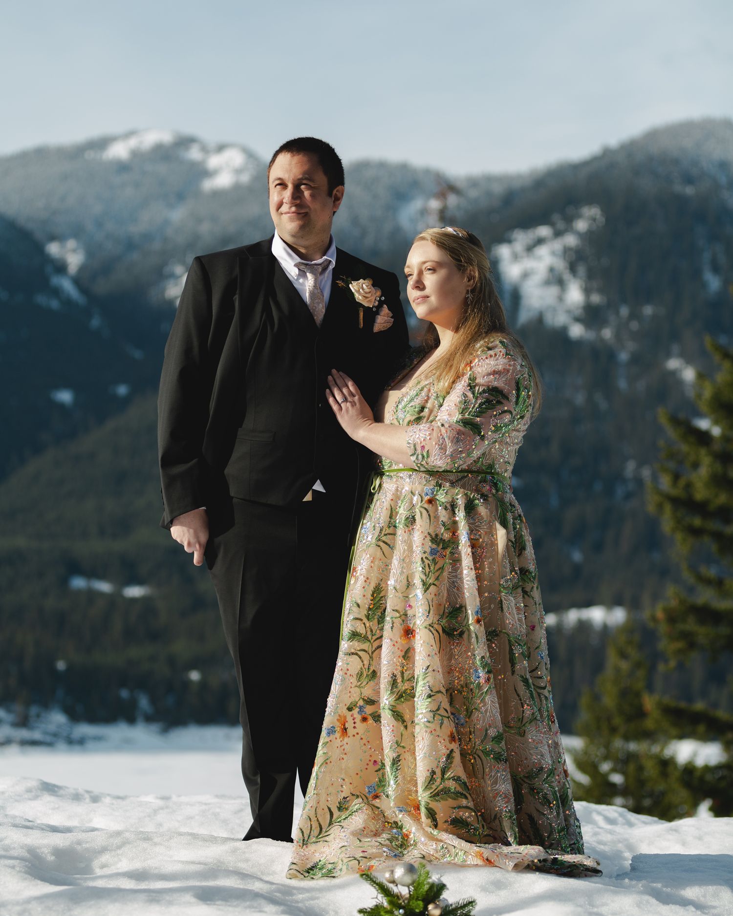 A couple in formal attire poses together on a snowy mountain landscape with evergreen trees and peaks in the background.