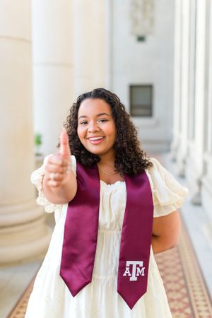 A graduate in a white dress and maroon Texas A&M stole gives an enthusiastic thumbs up while smiling on campus.