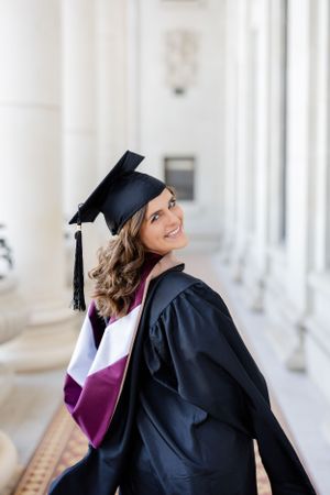 Graduate poses in black cap and gown with purple stole against white pillared building background.