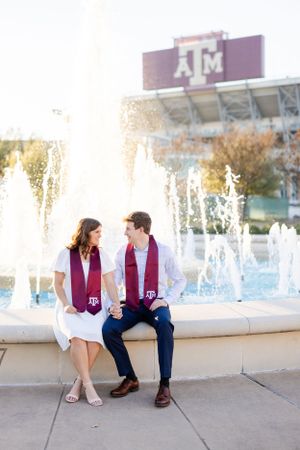 Students in maroon stoles sit together by fountains at Texas A&M University campus with iconic stadium sign in background.