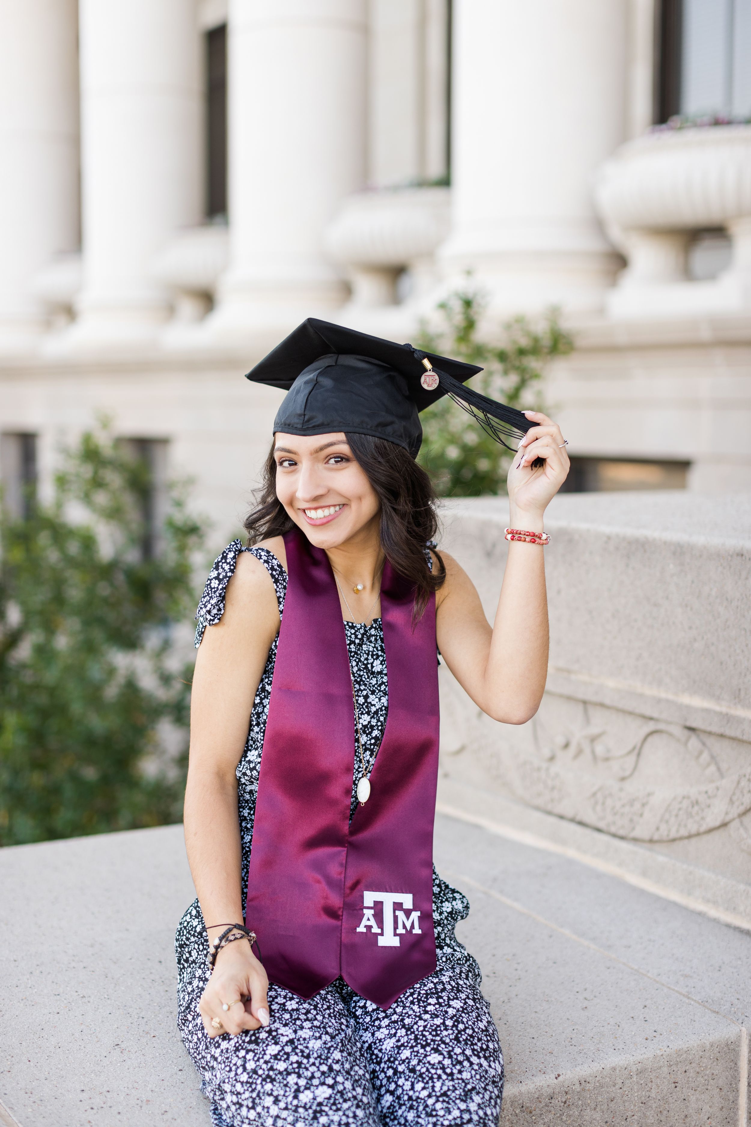 Graduate in maroon Texas A&M stole and graduation cap poses on campus steps wearing a floral pattern dress.