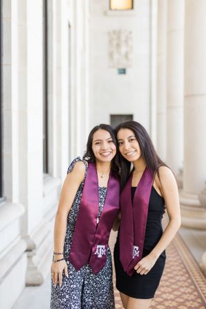 Two people wearing Texas A&M themed attire pose together in a bright hallway with classical white columns.