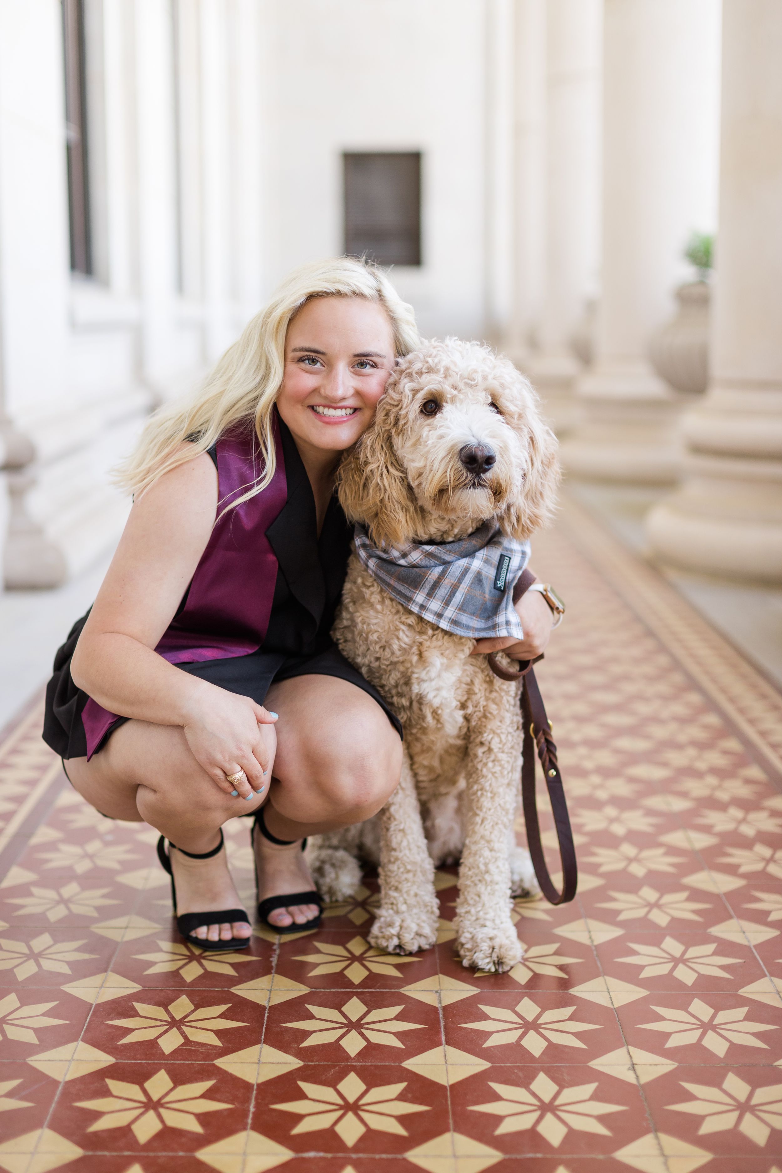 A goldendoodle dog wearing a plaid bandana poses with someone on an ornate tiled floor in an elegant hallway.