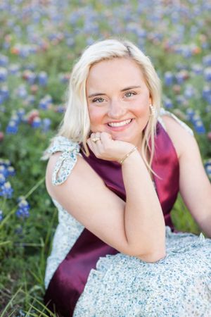 A senior portrait taken in a field of bluebonnets with a student wearing a maroon sash and floral dress.