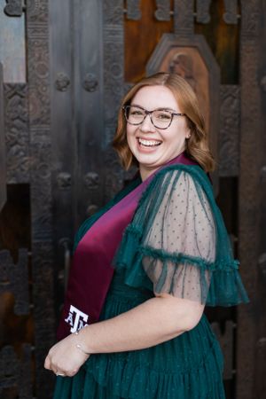 A joyful person in glasses wearing a burgundy sweater and emerald green dress with sheer sleeves poses against ornate doors.
