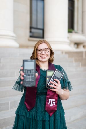 Student in green dress and Texas A&M sash holds up graphing calculators while smiling on campus steps.
