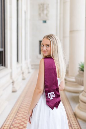 A graduate in a white dress and maroon Texas A&M stole stands in an elegant hallway with classical columns.