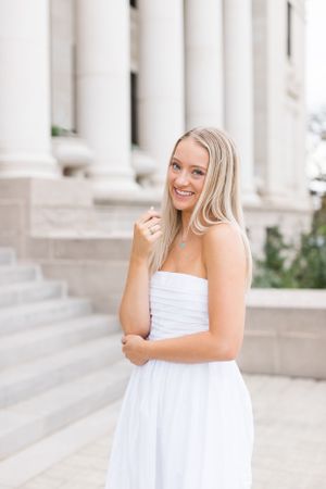 A person in a white strapless dress smiles while standing in front of classic stone columns on a sunny day.